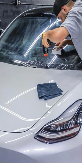 Technician using a heat gun and squeegee to apply window tint or protective film to the windshield of a white Tesla, with a microfiber cloth resting on the hood.
