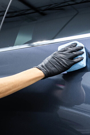 Gloved hand using a blue microfiber cloth to clean or polish the side door panel of a dark-colored car, likely during a detailing or ceramic coating process.