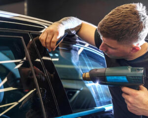 Male technician using a heat gun and squeegee tool to apply window tint film to a car door, ensuring a smooth and secure fit.