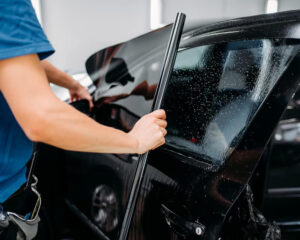 Auto technician preparing to apply a sheet of window tint film onto a wet car window, with water droplets visible on the glass during the installation process.