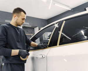 Auto detailer wearing gloves and AirPods applying masking tape around the windows and door frame of a white car, preparing it for tinting or paint protection work.