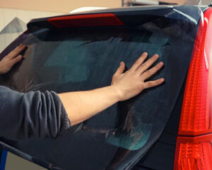 Person using both hands to press and smooth out window tint film on the rear windshield of a black SUV, with the film partially applied and visible wrinkles.