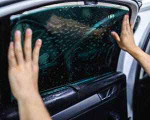 Person using both hands to press a tinted film onto the inside of a car's rear door window, with water droplets visible during the installation process.