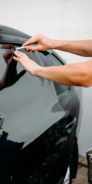 Technician applying window tint film to a black car's rear side window using a precision blade.
