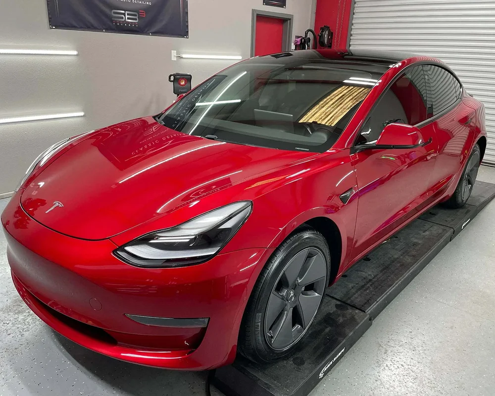 Bright red Tesla Model 3 with a glossy finish parked on a platform inside a detailing studio, reflecting overhead lights on its polished surface and showcasing black wheels and a glass roof.