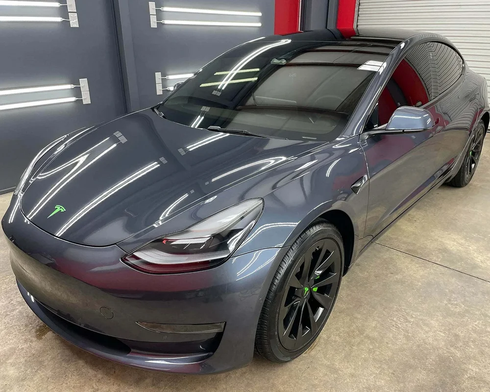 Glossy dark gray Tesla Model 3 with tinted windows and subtle green accents on the logo and wheel center caps, parked in a detailing shop with bright overhead lighting reflecting off the surface.