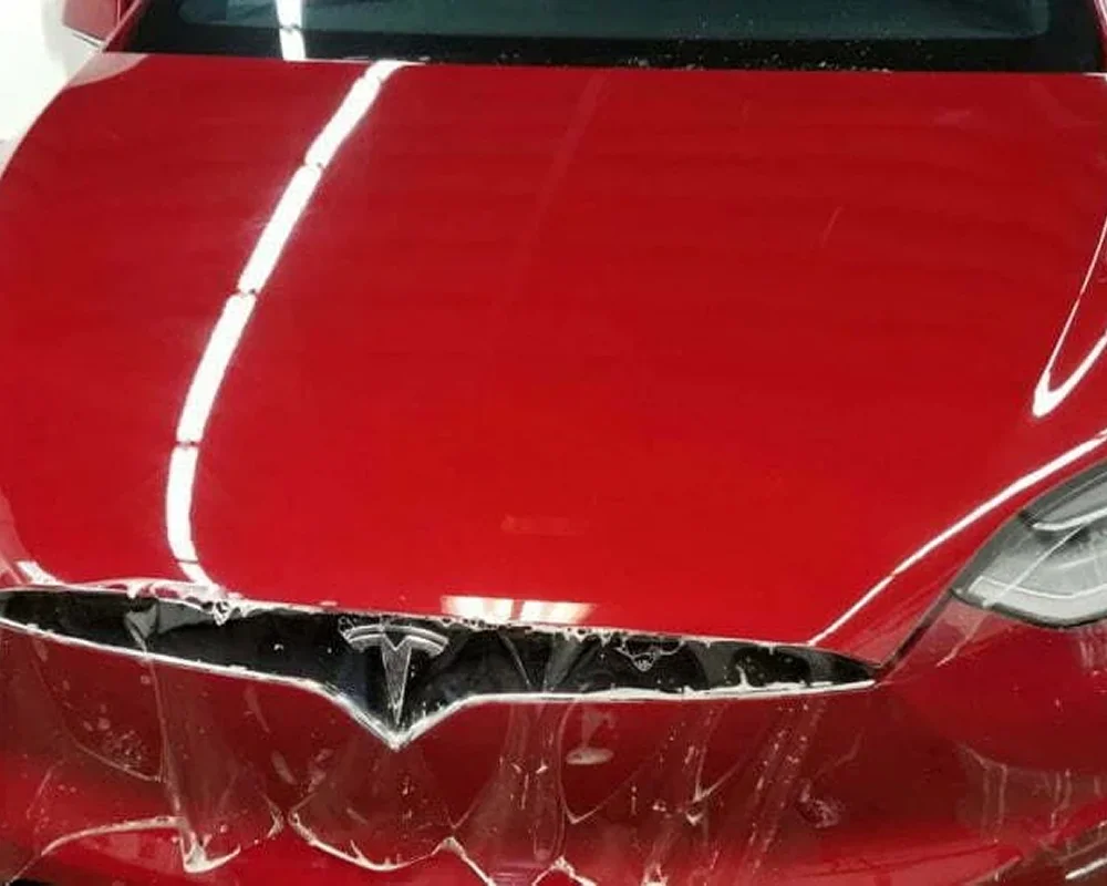 Close-up of the front hood of a red Tesla car with a protective clear film being partially peeled off, revealing the shiny surface underneath and the Tesla logo at the center.