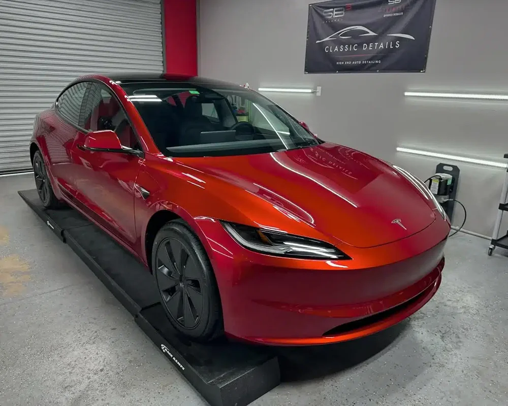 Shiny red Tesla Model 3 parked inside a detailing garage, elevated on a black platform with lighting reflecting off its smooth, freshly polished surface. A banner on the wall reads "Classic Details High End Auto Detailing."