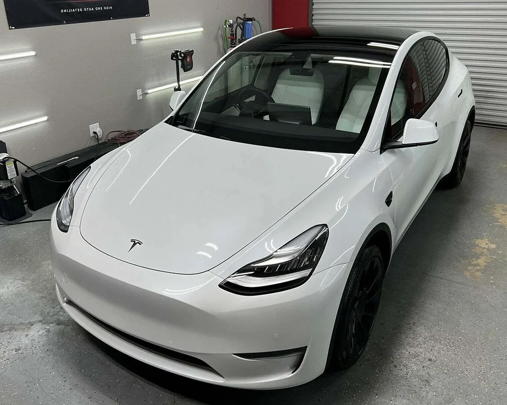 Glossy white Tesla Model Y parked inside a detailing garage with a clean, polished finish, black wheels, and a black glass roof under bright overhead lights.