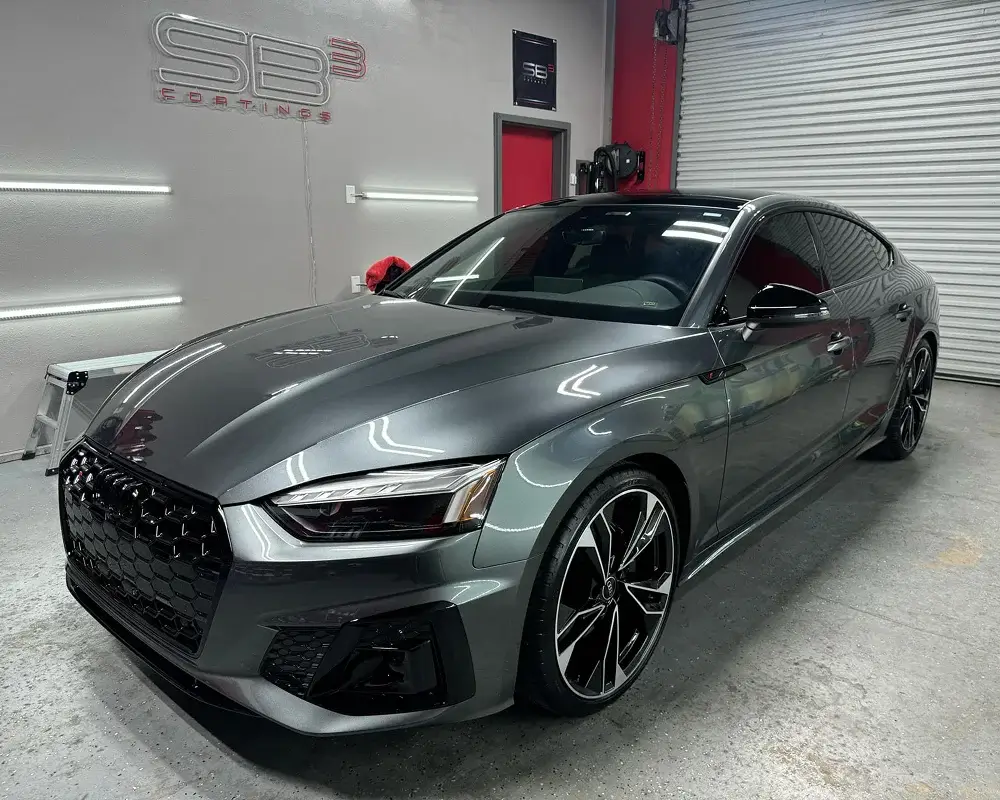 Metallic gray Audi S5 Sportback with glossy black accents and dark tinted windows, parked in a detailing studio under SB3 Coatings branding.