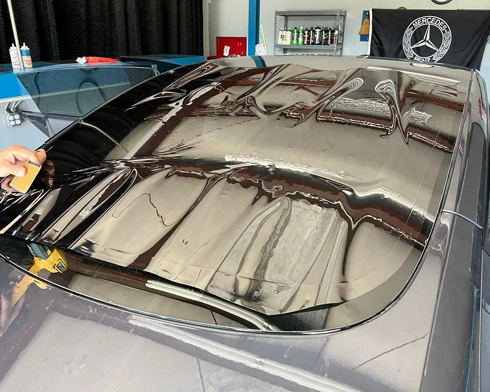 Close-up of a rear windshield as a technician applies dark window tint film using a squeegee, with visible air bubbles and reflections of the shop ceiling.