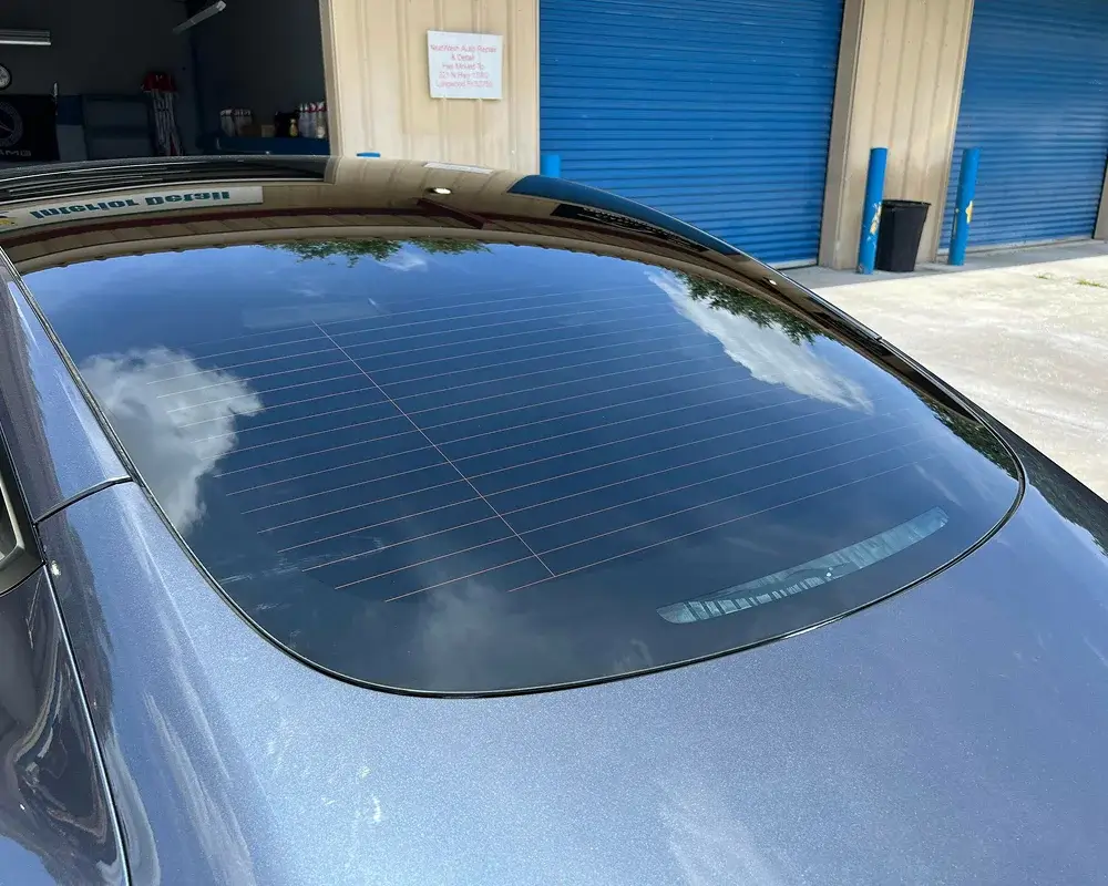 Rear window of a dark-colored car showing a clean, reflective surface with visible defroster lines, parked outside a building with blue roll-up doors and a repair shop sign.