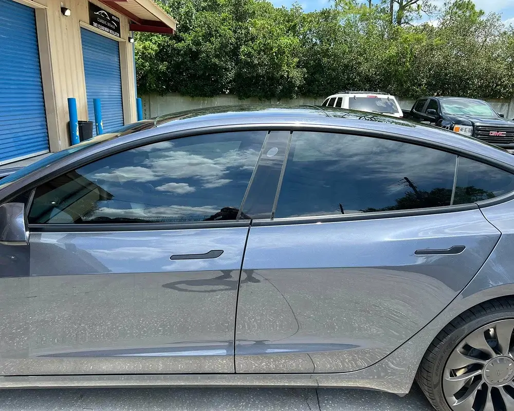 Side view of a sleek, dark gray Tesla Model 3 parked outside a building with blue storage doors, reflecting clouds and sky on its tinted windows.