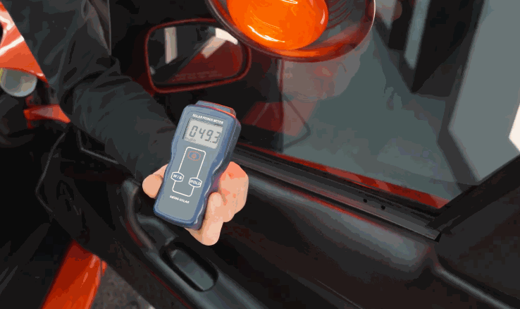 A technician holds a digital tint meter against a car window, displaying a visible light transmission reading of 49.3 percent. The car’s side mirror and part of the red exterior are visible in the background.