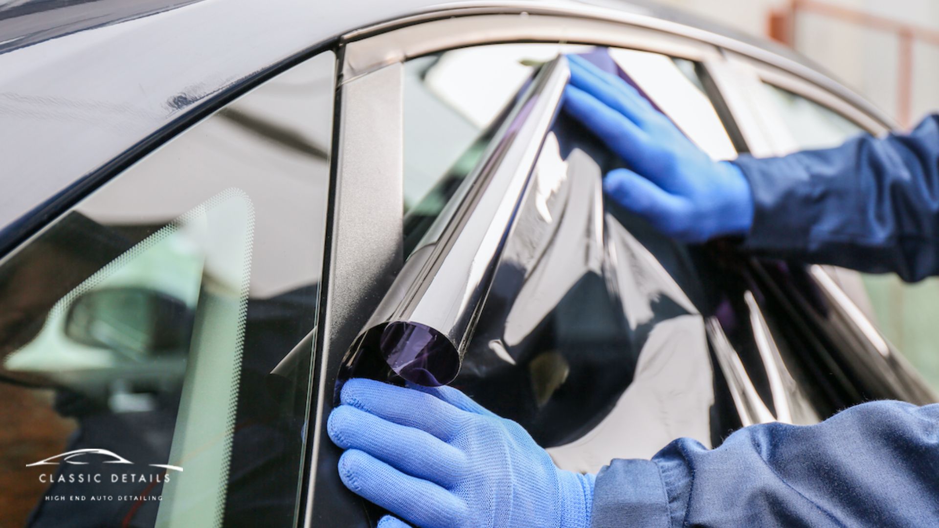 A person in blue gloves installs a roll of dark window tint film onto a car window, smoothing it out with both hands.
