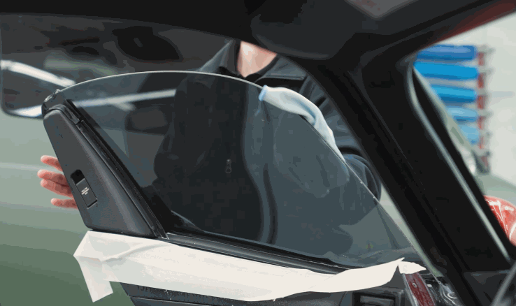 A person carefully aligns and applies a sheet of window tint film to a clean car door window, using masking tape to protect the car’s interior trim.