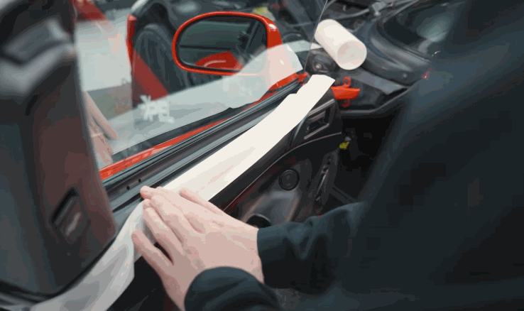 Close-up of hands pressing down on tape securing a car window's edge, preparing the area for tint installation by protecting the trim.