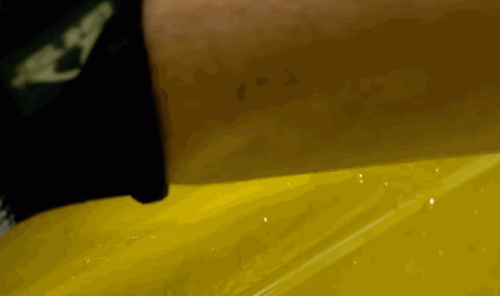 A close-up of a hand smoothing down paint protection film on a bright yellow car surface, with small bubbles visible underneath.