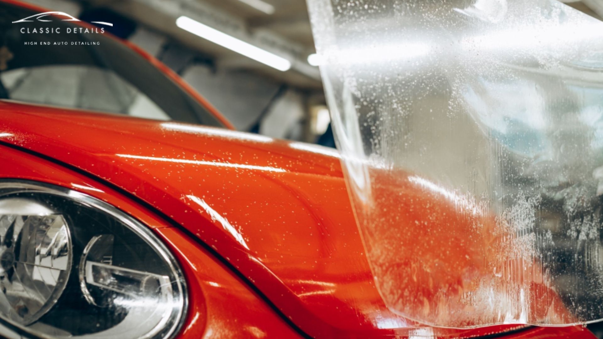 A close-up of a red car's front end as a transparent paint protection film is installed, with visible water droplets between the film and paint.