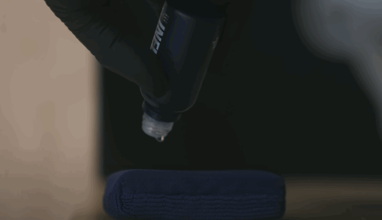 A close-up of a drop of ceramic coating liquid being applied to a microfiber applicator pad, held by a gloved hand in a dimly lit workspace.