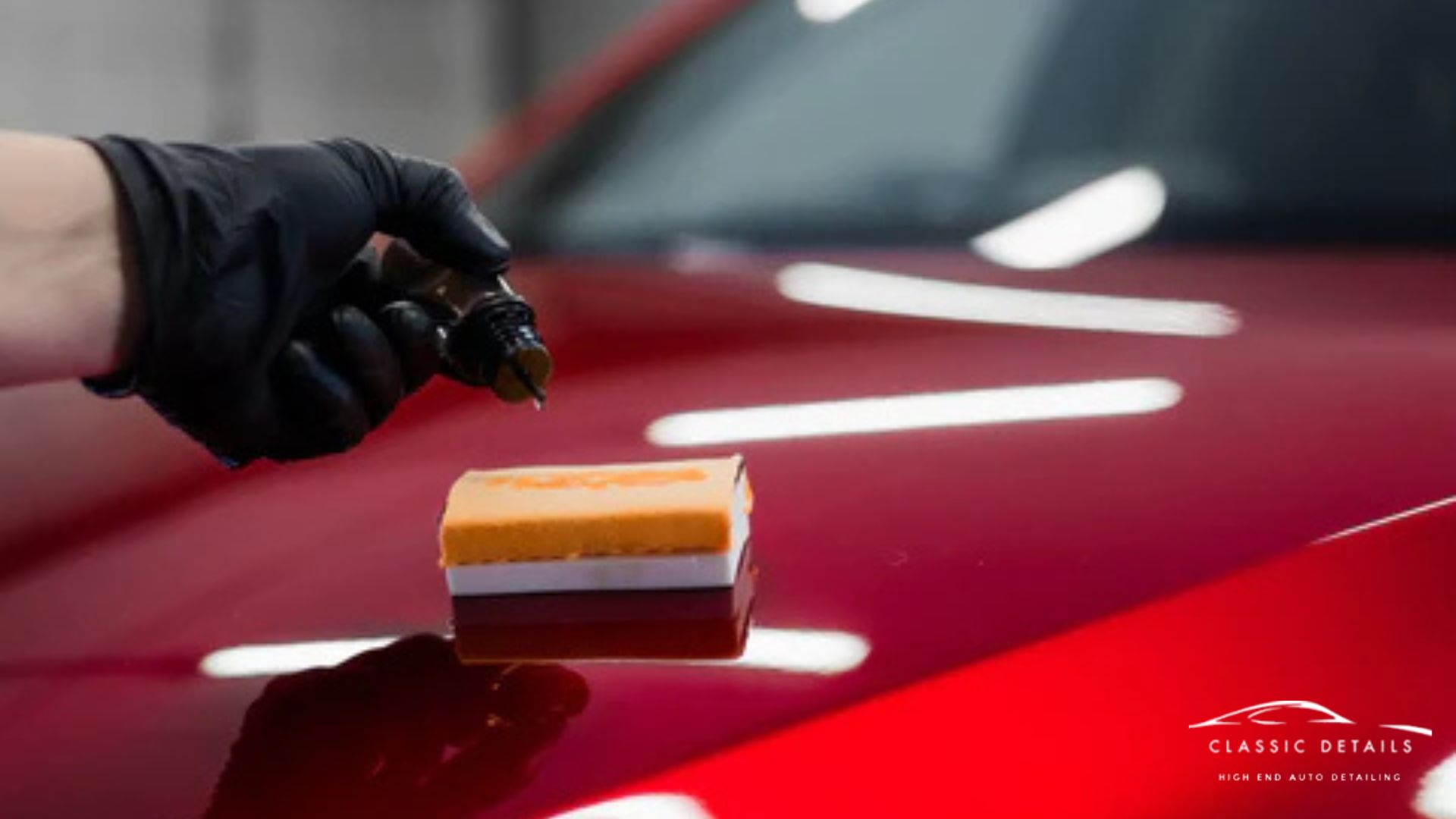 A detailer wearing a black glove applies ceramic coating to an applicator sponge over the glossy red hood of a car