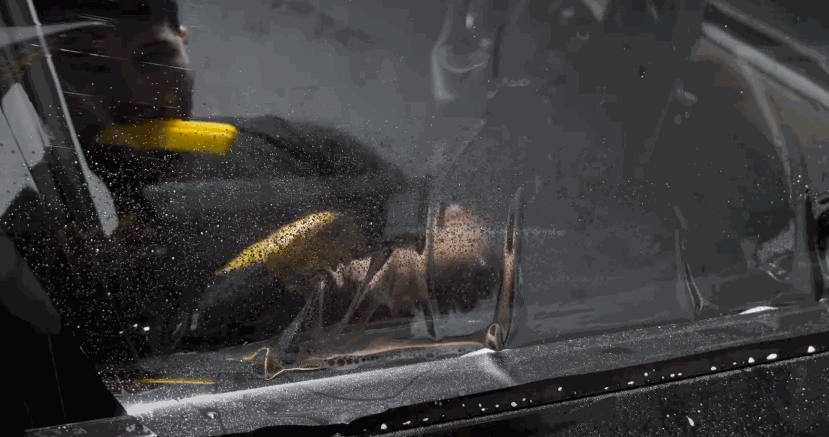 Technician using a yellow squeegee to apply car window tint film to a wet car window. Water droplets and air bubbles are visible under the film.