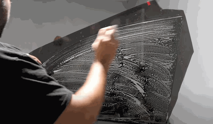 A person cleaning a car window with soapy water in preparation for car window tint installation. The surface is covered in soap streaks and foam.