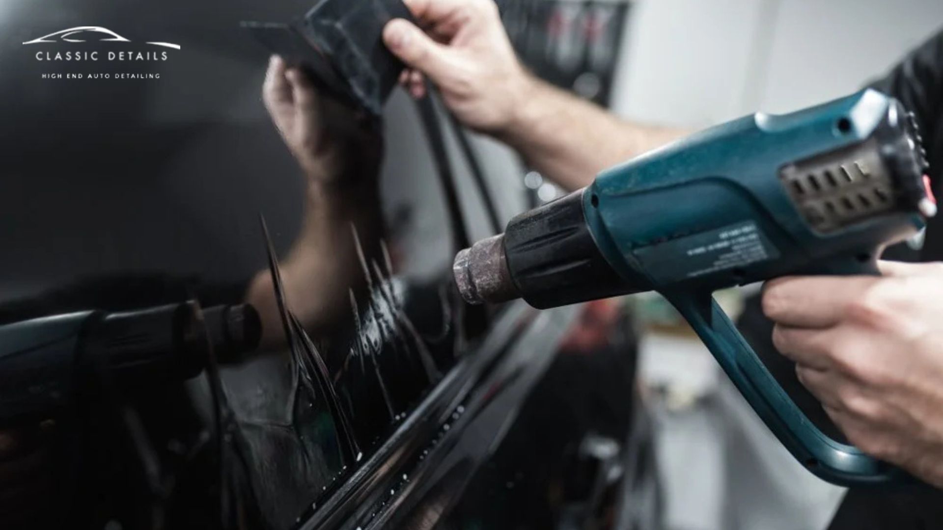 A heat gun is used to shrink and mold the car window tint film onto the window while another hand smooths it out with a hard tool.