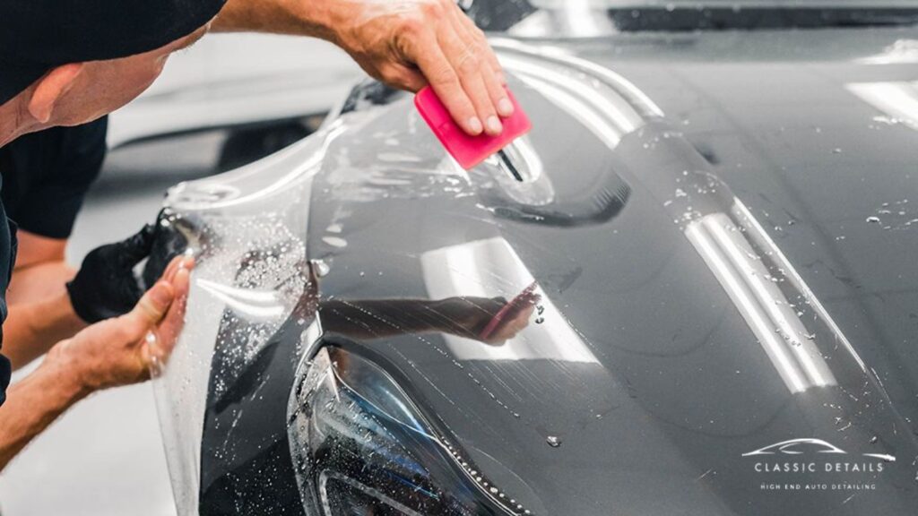 Technician using a pink squeegee to smooth out PPF on a wet car hood while aligning the film over curves and edges.