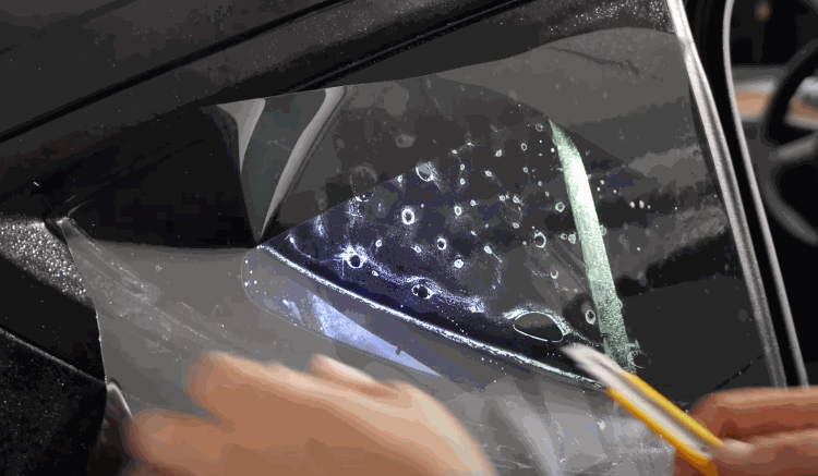 Installer trims and smooths window tint film onto a small triangular car window while water bubbles are visible beneath the surface. The process highlights detailed window tint work on tight corners and edges.