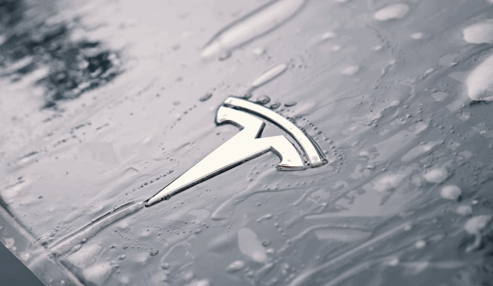 Close up of a Tesla emblem on a gray hood covered in water droplets, highlighting how moisture beads on the surface of the Tesla paint. The droplets reflect light, emphasizing the condition and protection of the finish.