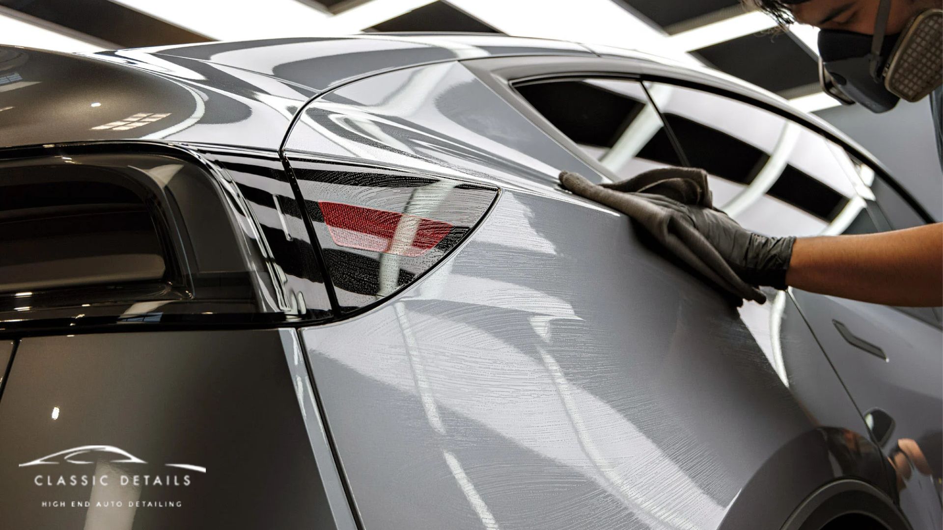 A technician wearing gloves carefully wipes the rear panel of a silver Tesla in a bright detailing studio, showing swirl marks being corrected in the paint.