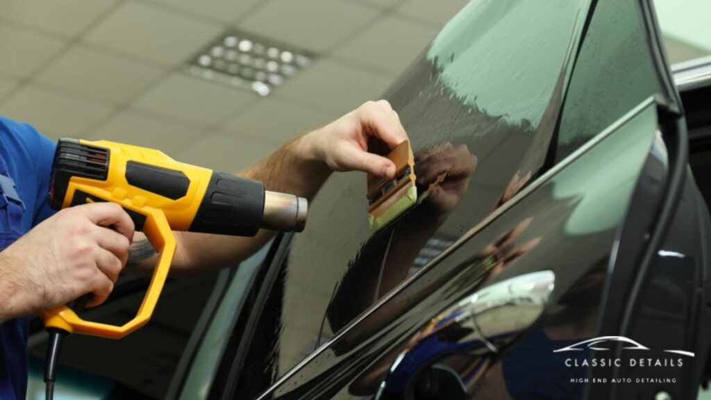 Technician uses a heat gun and squeegee to apply ceramic window tint to a black car door inside a detailing shop. The glossy surface reflects the worker’s hands and tools