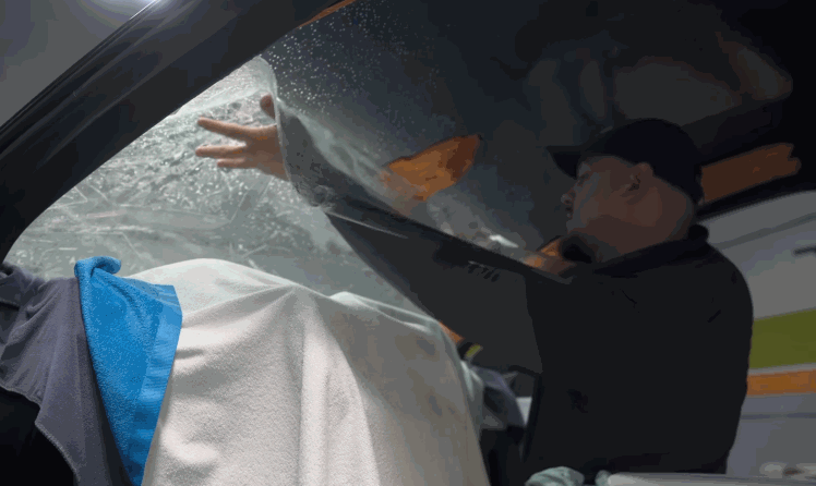 Installer carefully smooths ceramic window tint film onto a car window from the inside while water droplets and a towel are visible during the process. The film is being aligned and pressed flat to ensure a clean bubble free result.