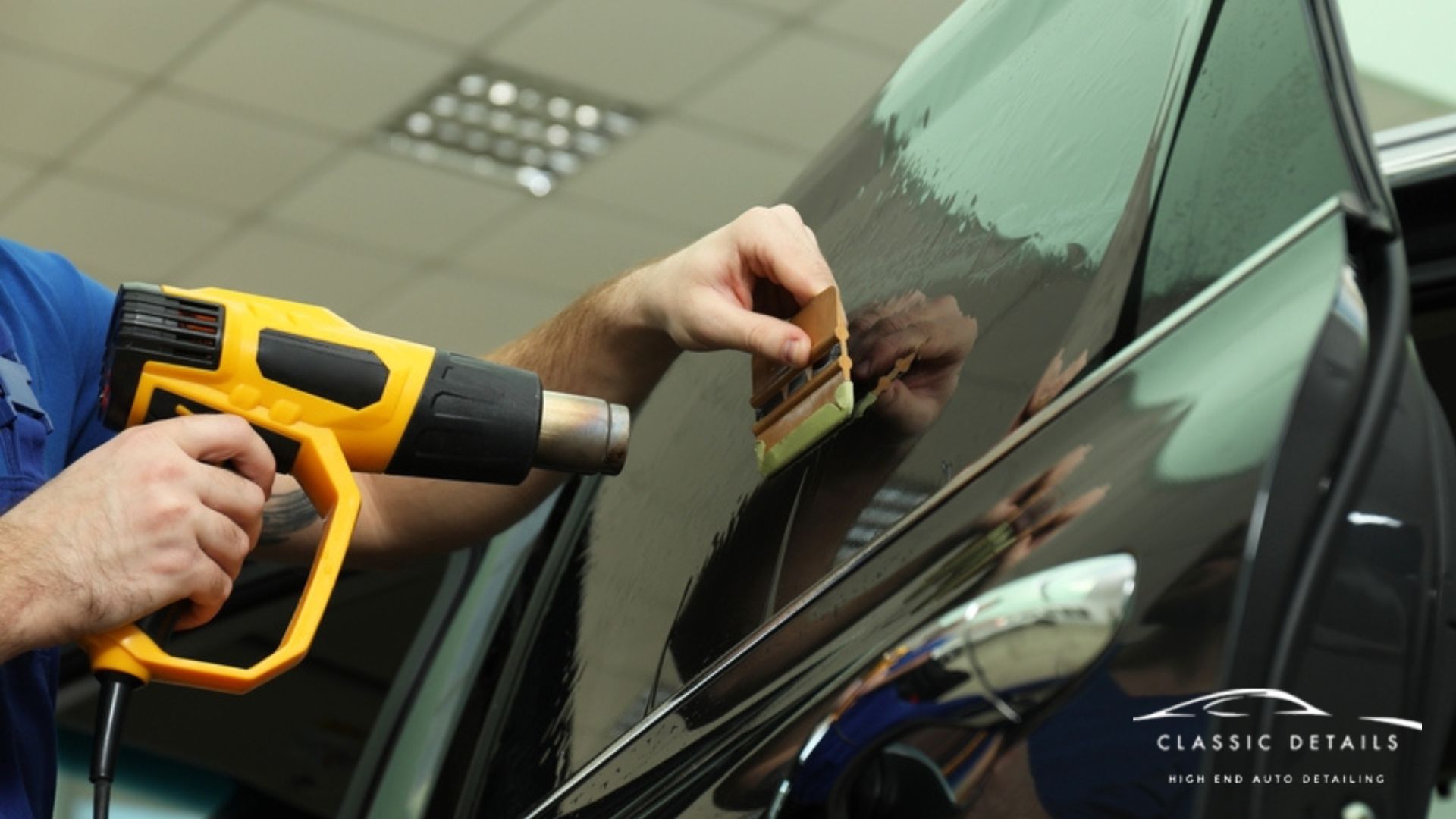 Technician uses a heat gun and squeegee to apply ceramic window tint to a black car door inside a detailing shop. The glossy surface reflects the worker’s hands and tools