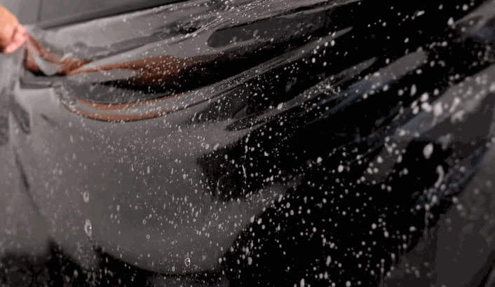 A sheet of protective film is being applied over a wet black Tesla surface as water sprays across the panel during installation. The process demonstrates how Tesla paint can be shielded from damage and harsh environmental exposure.