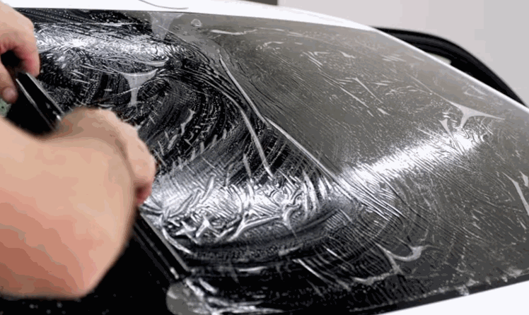 Close up of hands using a squeegee to remove solution and air bubbles from ceramic window tint film on a car window. The wet surface shows streaks being pushed out as the tint adheres smoothly.