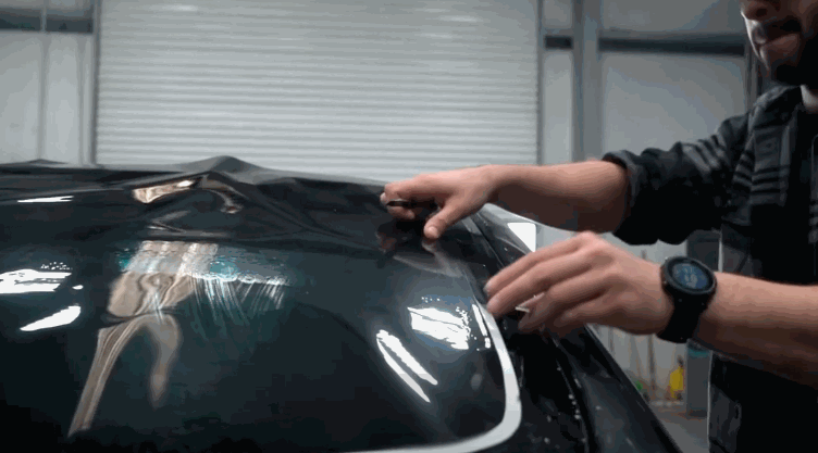 Technician carefully applies window tint film to a dark car window inside a detailing shop. Overhead lights reflect across the glass as the film is positioned by hand.