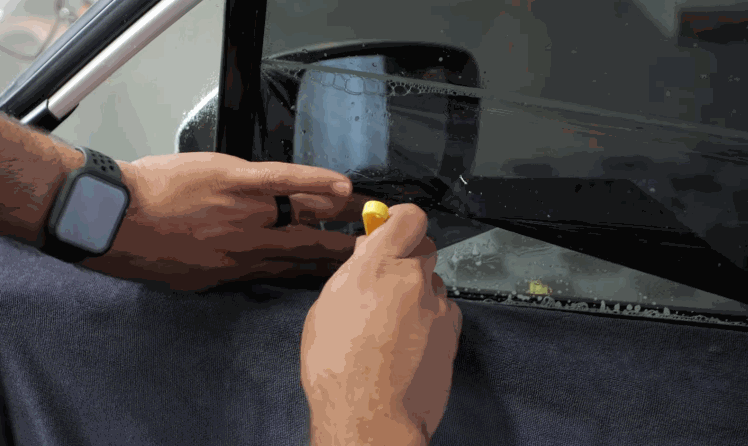 A technician trims wet window tint film along the edge of a car side window. Close hands and tools show the precision needed for a clean fit.