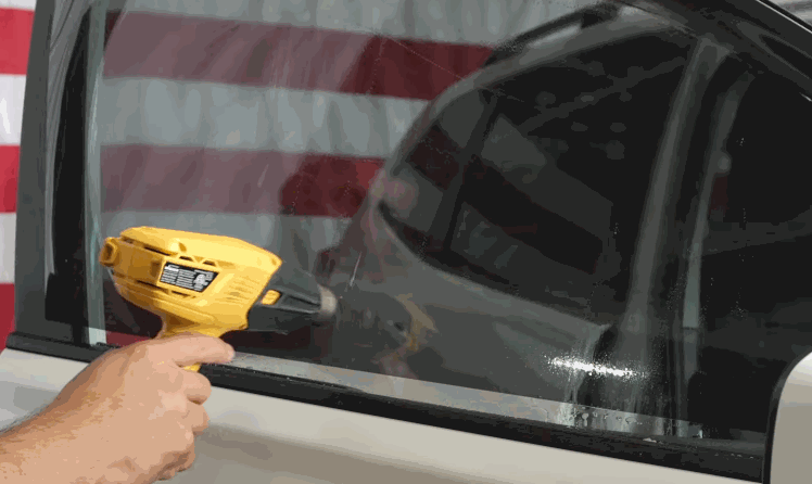 A technician uses a yellow heat gun on a dark car side window while shaping window tint film. The American flag reflection in the glass adds a bold backdrop to the installation.