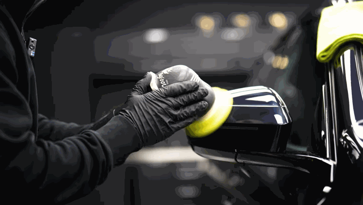 A technician hand polishes the side mirror of a black vehicle before ceramic coating. The close up shows careful detailing around the mirror and trim.