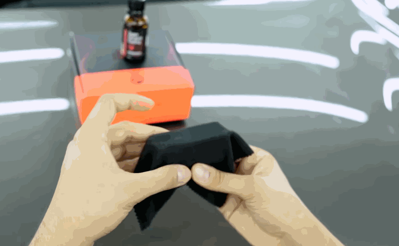Hands hold a black applicator block over a glossy vehicle hood before ceramic coating is applied.