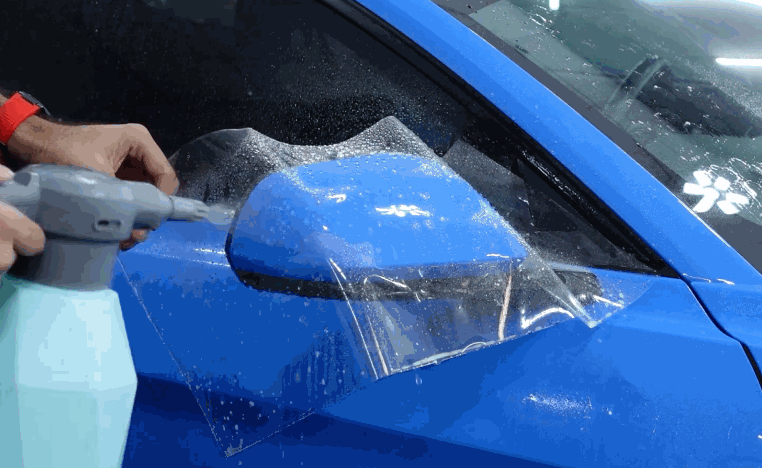 Technician sprays and positions paint protection film over the side mirror of a blue car.