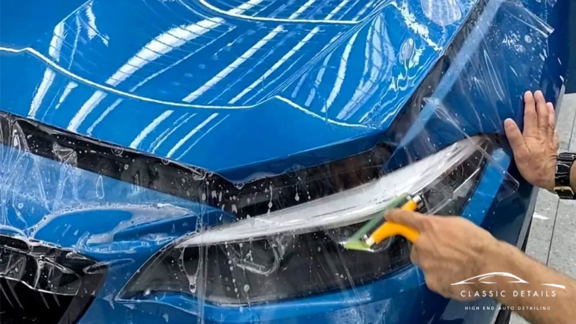 Technician applies paint protection film to the front headlight and fender of a blue car using a squeegee.