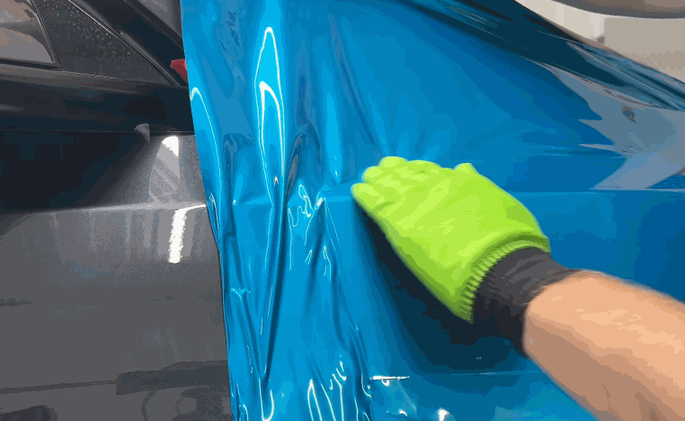 Gloved hand presses colored PPF across a bright blue car panel to smooth the film into place.