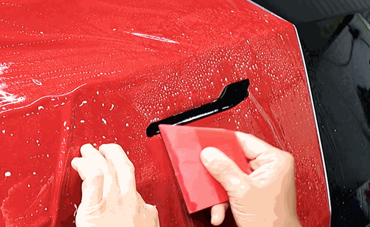 Installer smooths colored PPF across a red car panel with a red squeegee near a small trim opening.