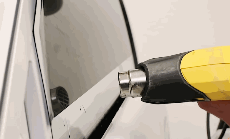 Heat gun warms window tint along the edge of a white car window during installation.