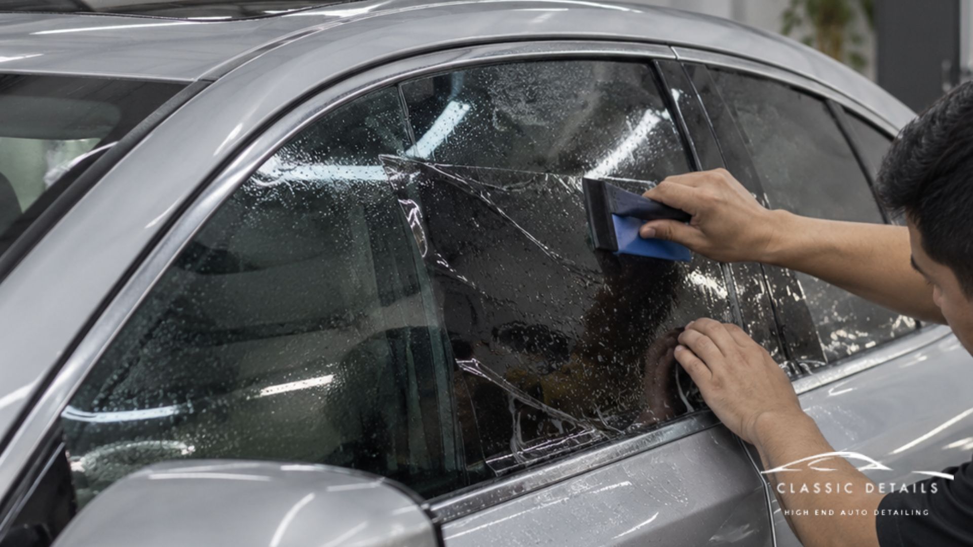 Installer smooths window tint onto the rear side window of a silver car using a squeegee.