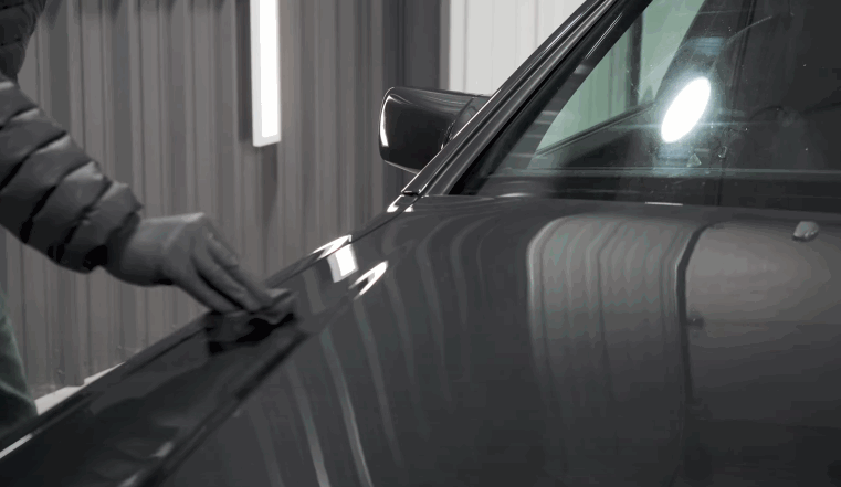 Technician wiping a dark vehicle hood with an applicator pad during a ceramic coating application in an indoor detailing studio.